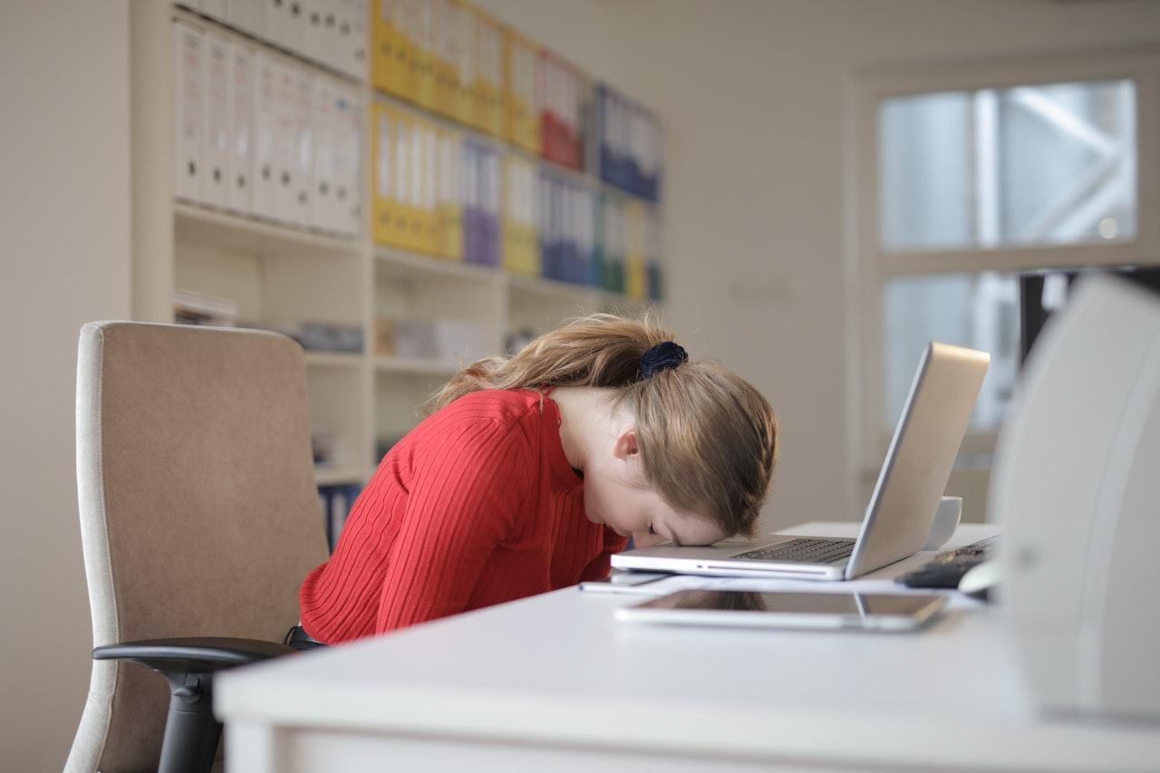 La imagen muestra a una mujer blanca con cabello rubio frente a un escritorio de oficina. Tiene la cabeza en un computador portátil y los ojos cerrados, por lo que se ve muy cansada.