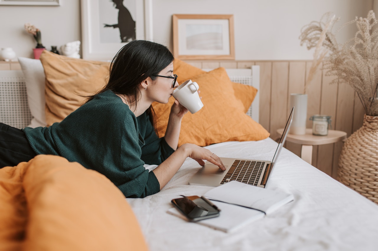 A woman lying on her bed, drinking coffee, and looking at her laptop