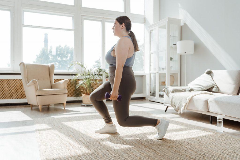 A woman exercising in her living room by holding dumbbells in her hands and stepping forward.