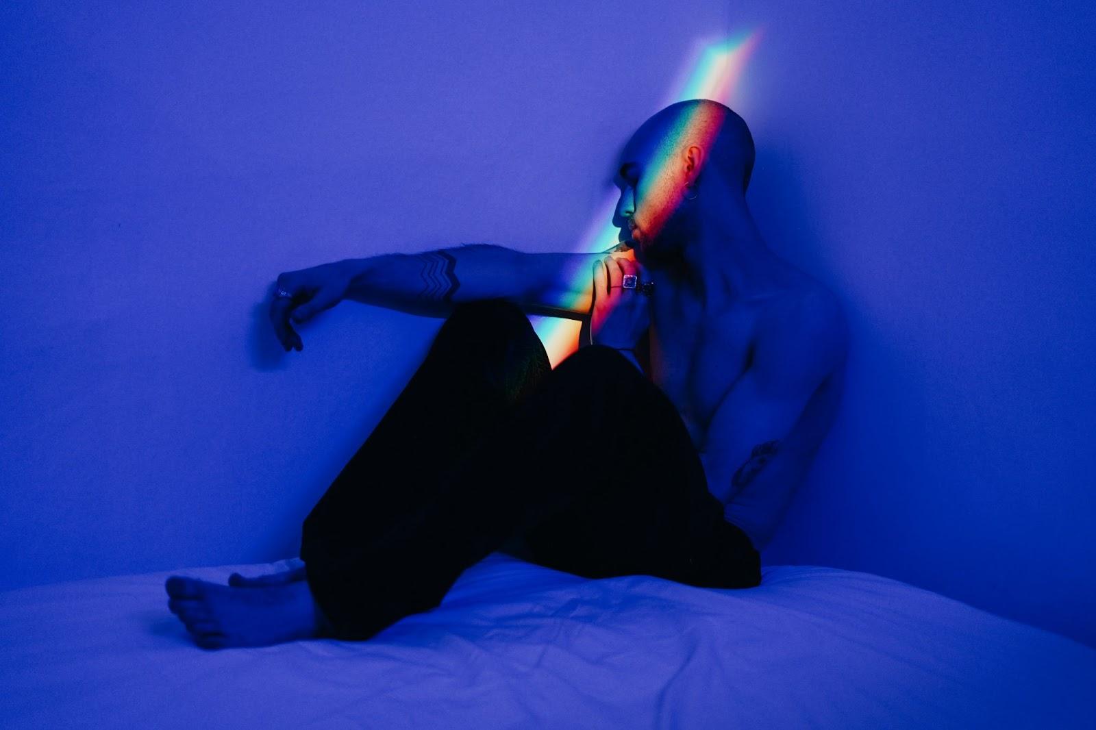 A man sits in a dark room, highlighted by a glimmer of rainbow light