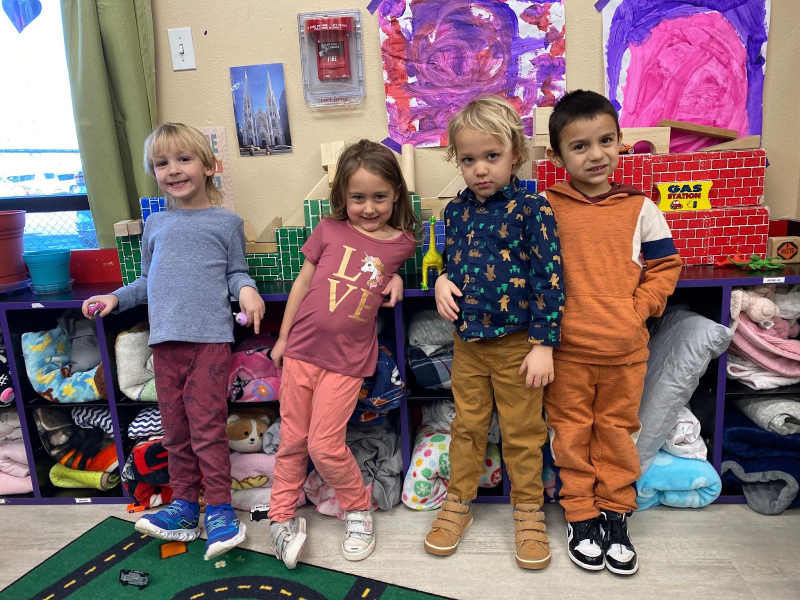 Four preschoolers smile at the camera while standing in front of artwork and naptime supplies.