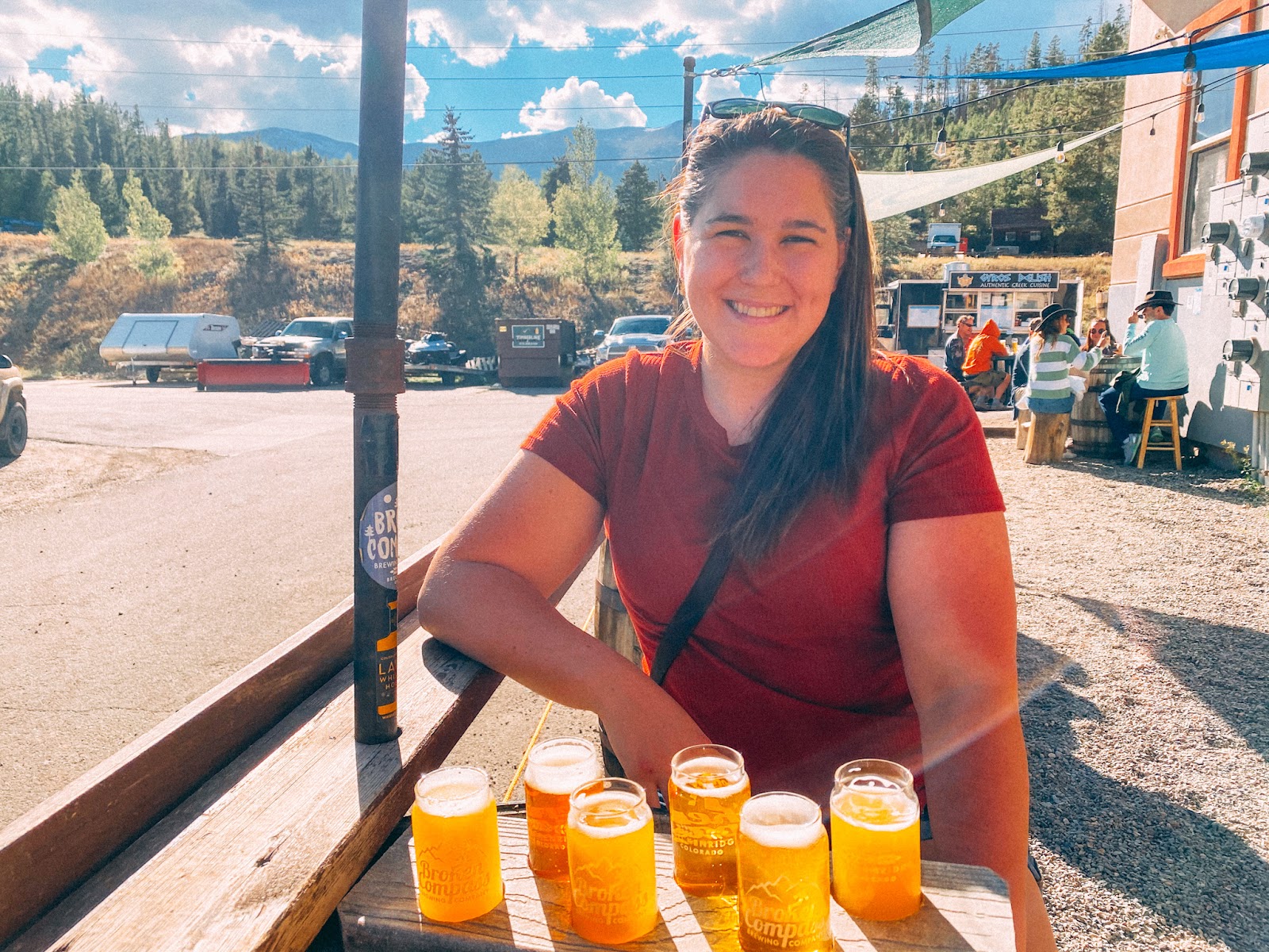 Allie from the Hoppy Passport is sitting with bottles of beer at an outdoor stand. Behind her is the mountain range and forest.