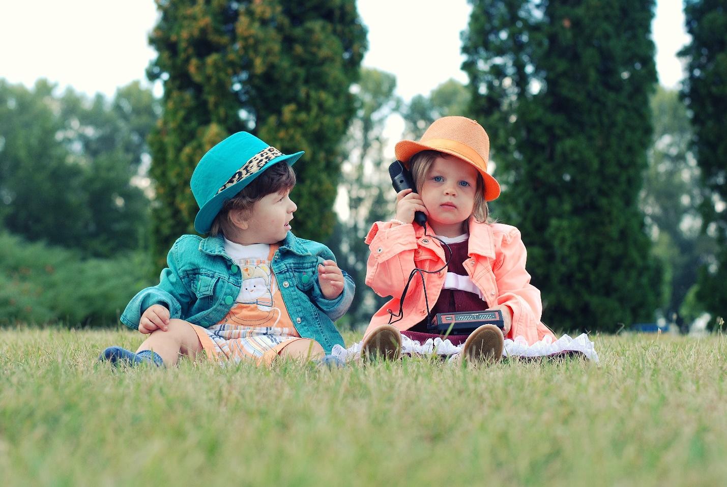 due bambine vestite con un vestito e un cappello colorato sono sedute sull'erba e una le tiene il telefono all'orecchio