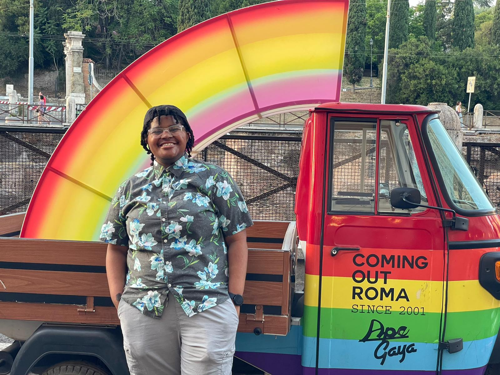 A person standing beside a pickup truck. The truck has a rainbow in its cargo area, and the front of the truck, painted rainbow, reads "Coming Out Roma".