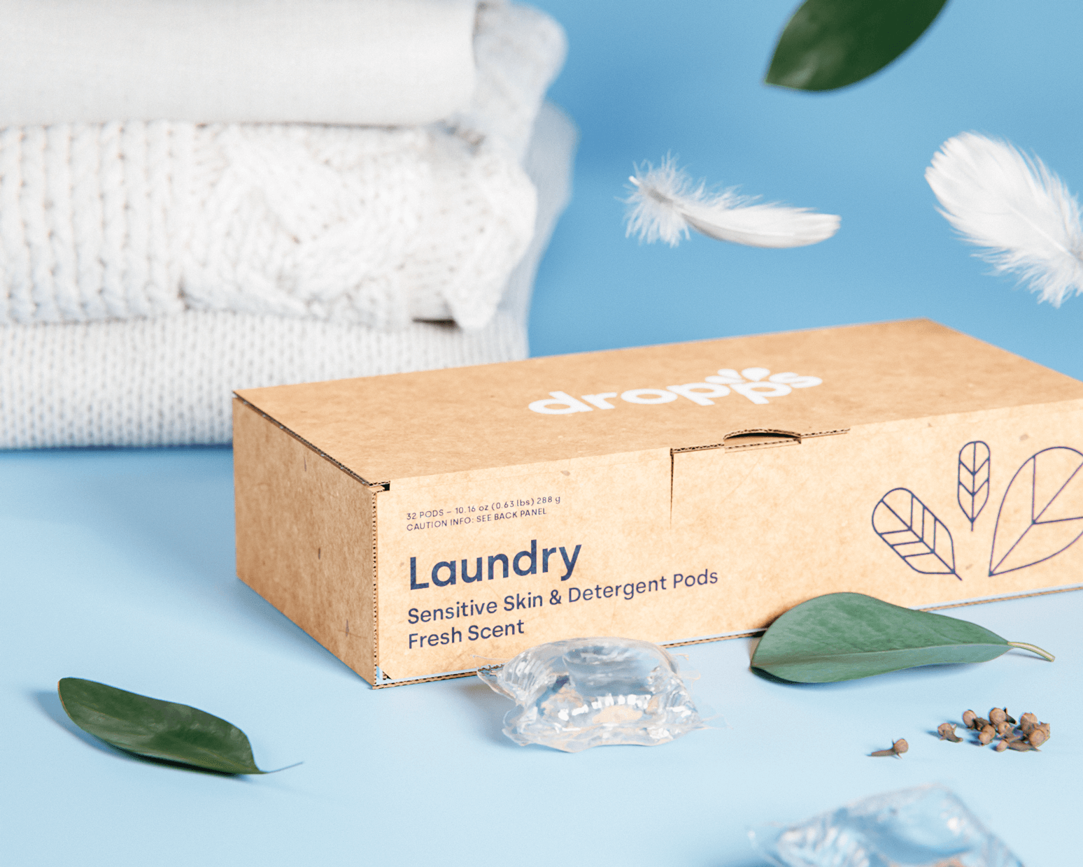Feeling Fresh and Cleaning Up the Laundry Industry with Dropps–A box of Dropps’ Sensitive Skin and Detergent Pods surrounded by white folded sweaters, feathers, leaves, and laundry pods. 
