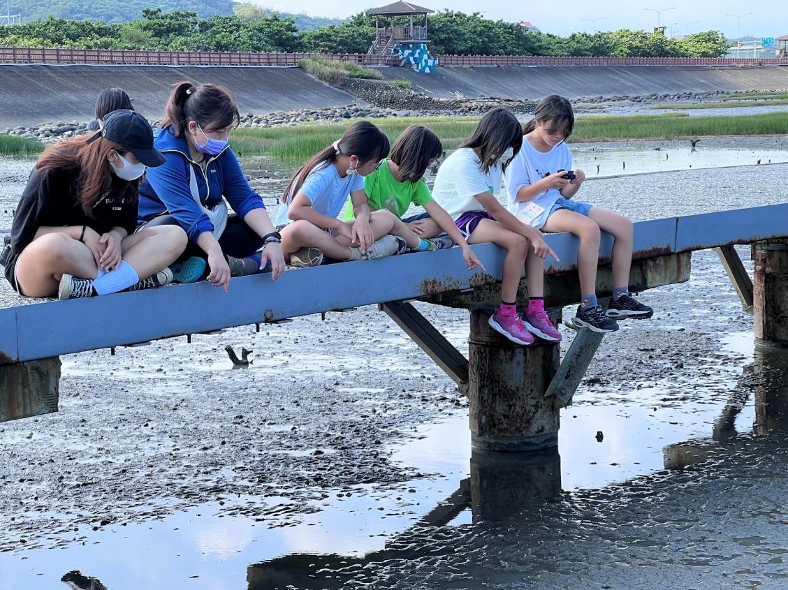 坐在河堤上，活力少女們跟著老師和志工在緊鑼密鼓的行程當中休息；她們指向地上的天空，因為水面映照著讓夢想飛得更遠的藍天。