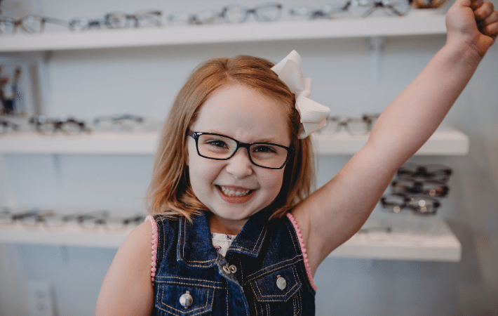 A little girl with red hair celebrating her first pair of glasses with her fist in the air from excitement.