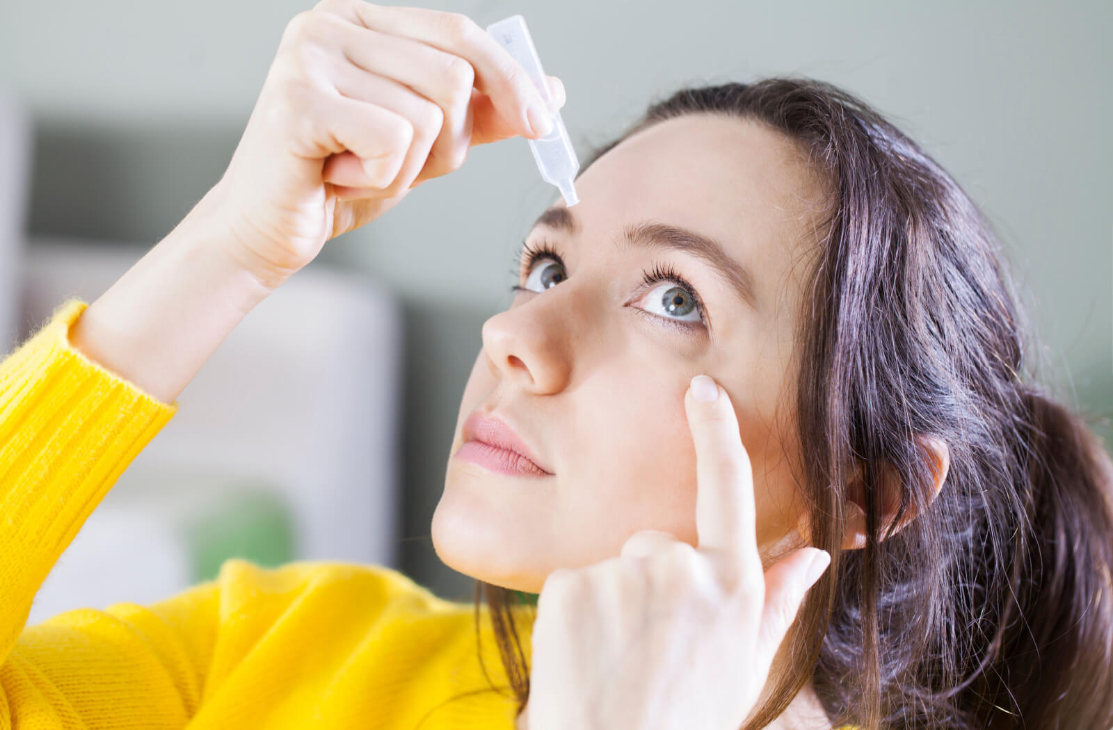 A woman holding a small bottle of eye drops in her right hand and putting them on her left eye while she uses her left index finger to pull her eyelid down.