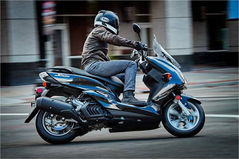 A Person on a Blue and Black Yamaha SMAX Scooter from their 2019 lineup. They are driving on a city street appearing to turn left. 