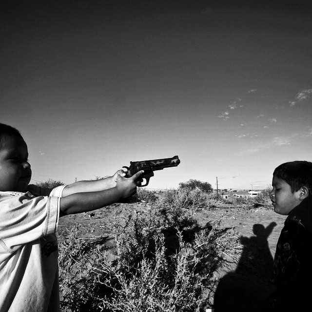 Dois meninos, de idade aparentemente próxima, em um lugar que parece ser um campo com arbustos. O menino da esquerda aponta uma arma para a cabeça do outro, que está de braços abertos e olhos fechados.