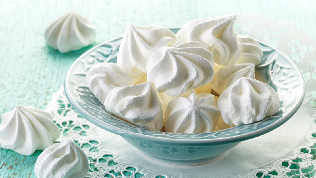 An aqua bowl filled with white meringues. The bowl sits on a lace napkin and a three white meringues are scattered around the bowl.  