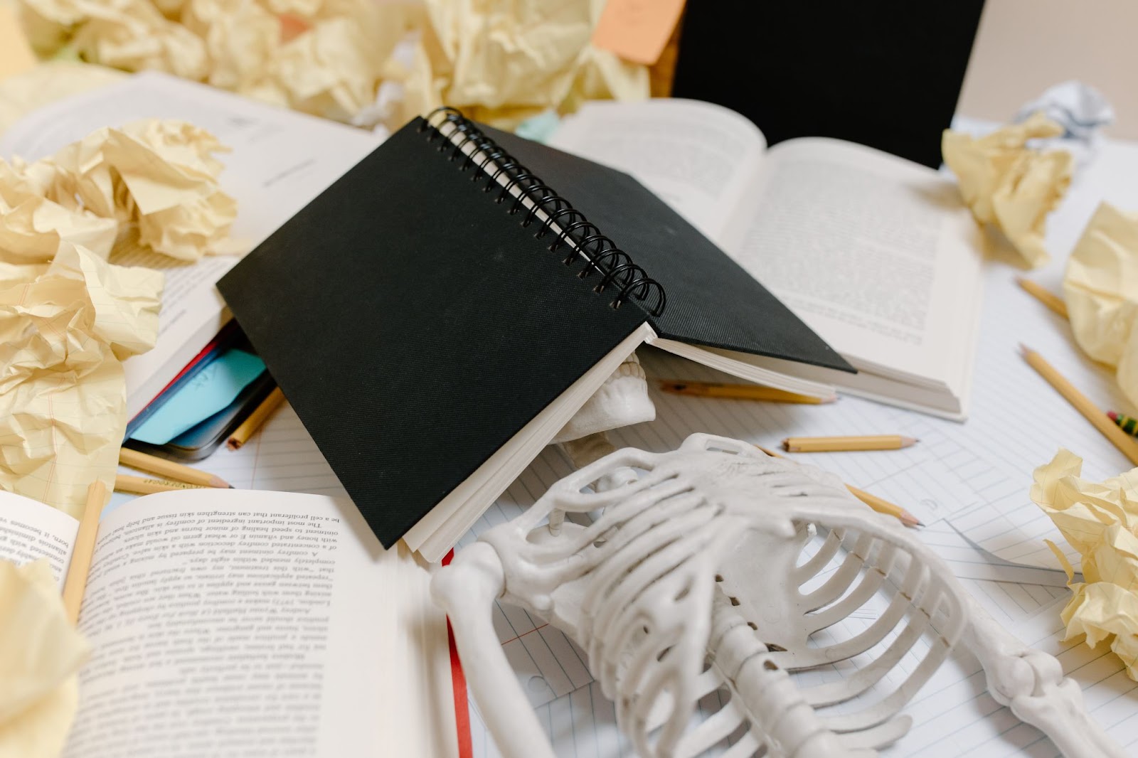 skeleton with book over its head, surrounded by books and crumpled up papers