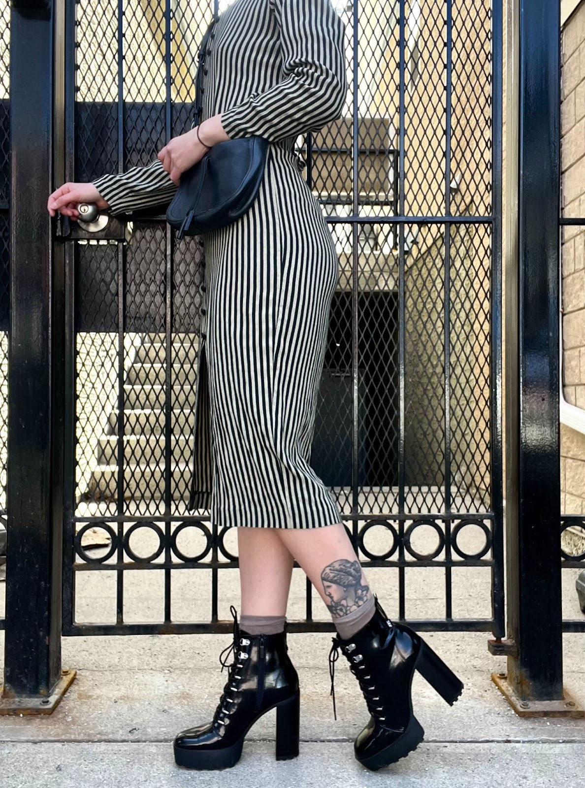 an image of a woman from the shoulders down. She is turned to the side in front of a metal door. she is wearing a long sleeve striped dress, and has accessorized with a black purse, belt and high heeled boots 