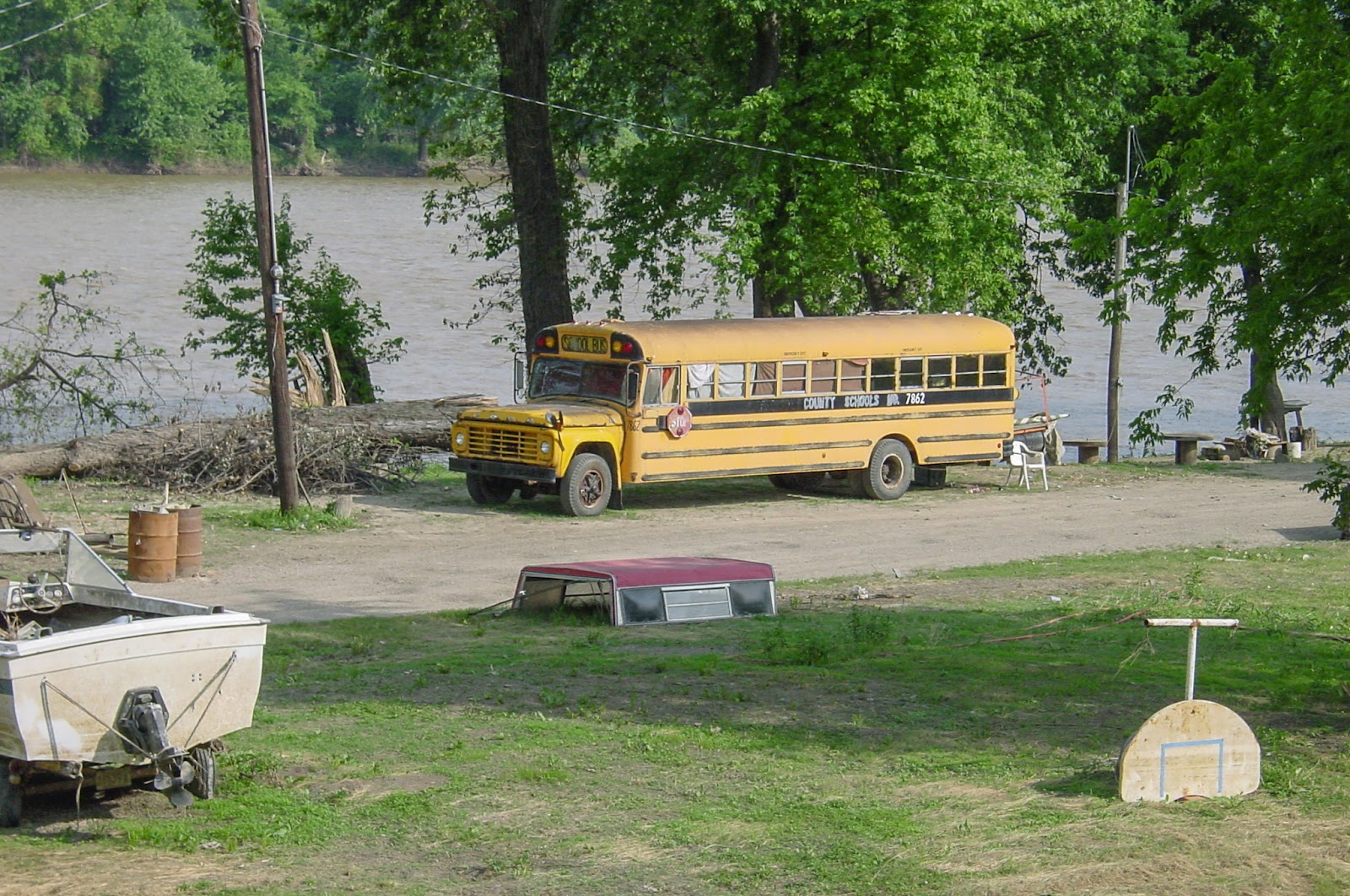 A boat and school bus altered to be living quarters sit on the edge of a river with a broken basketball backboard in mowed grass. 
