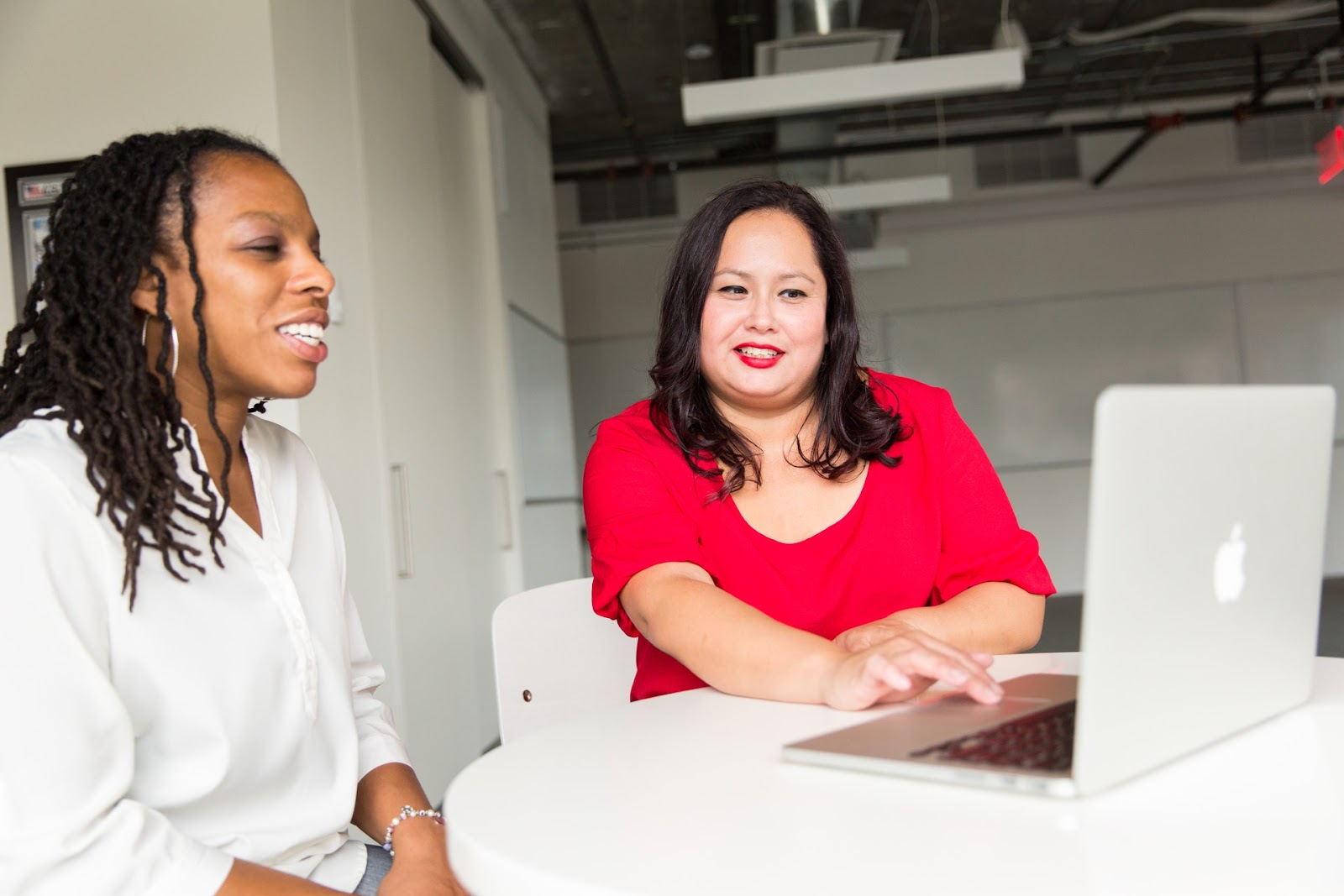 Two adults, one in a white shirt and one in a red shirt, are seated at a table. They smile as they discuss something they are both viewing on the laptop screen on the white table in front of them. They appear happy.