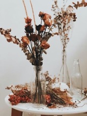 A tall bouquet of red and pink fried flowers in the foreground on a white table with other dried flowers at the base. In the background are two other vases, one with flowers, one without.