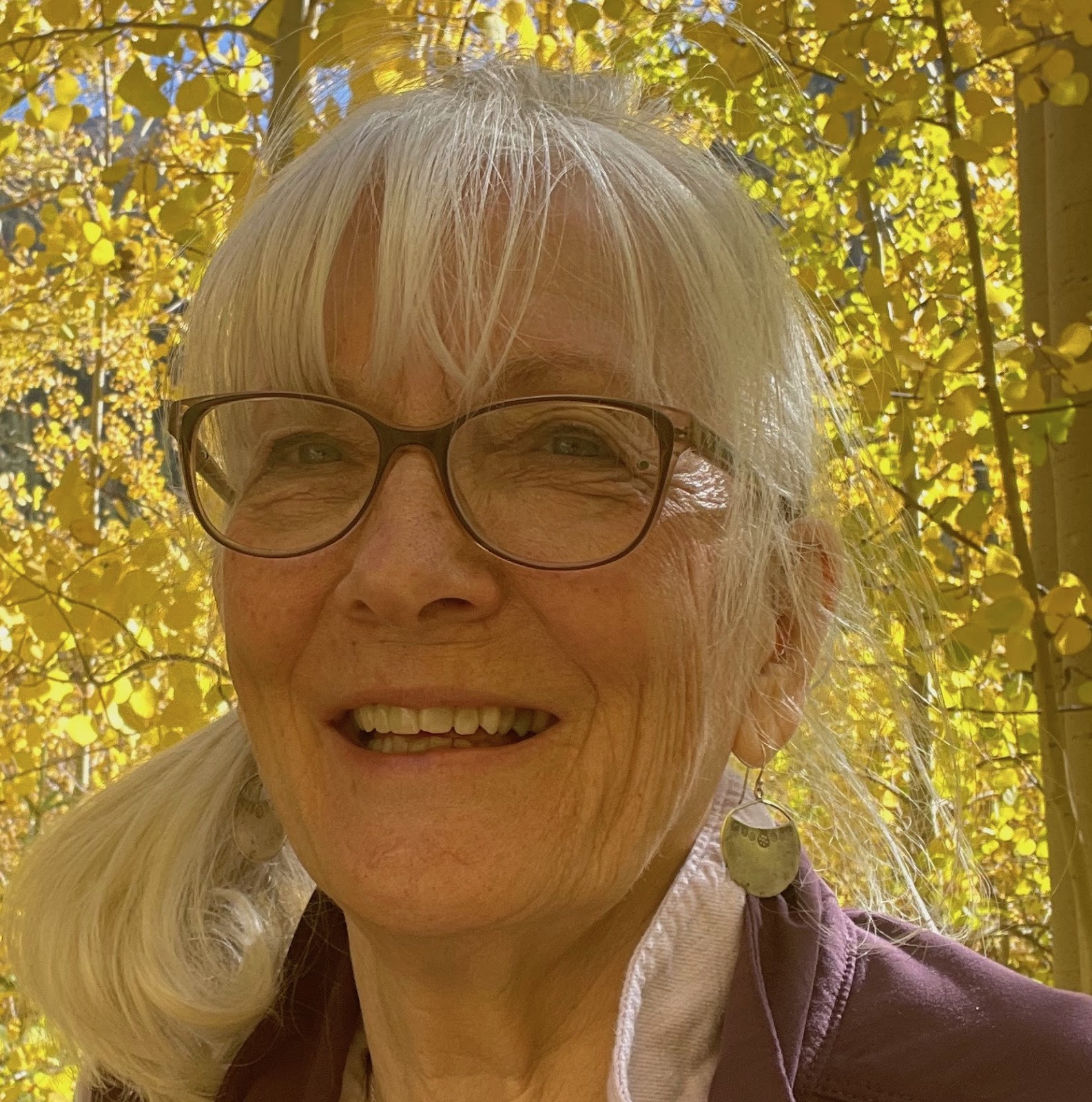A woman's face is framed by yellow aspen trees. She is smiling and wears glasses.