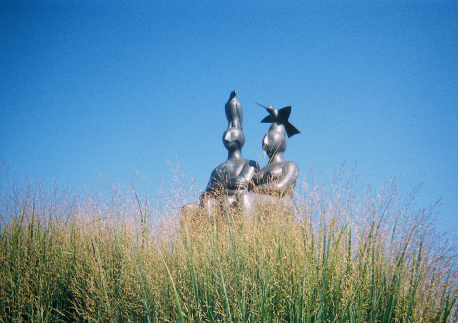 Image: Distance shot of Serenity, 2005. Green grass in the foreground partially covers a sculpture of two figures against a blue, cloudless sky. Photo by the author.