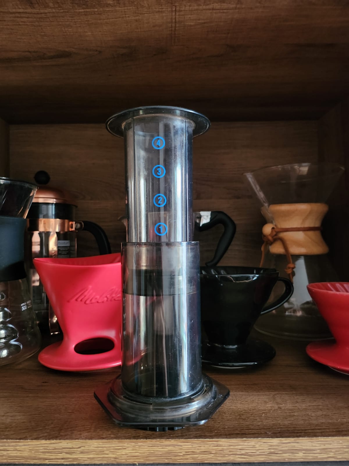 Foto deaeropress, feito em plástico, na cor preta transparente.
Imagem com filtro Aeropress em destaque, com fundo de madeira. Outros métodos diversos de café ao fundo, desfocados.