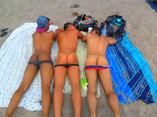 three gay males at the beach lying down on their stomachs on their beach towels with their speedo swimwear pulled down to revealed their hairless ass cheeks