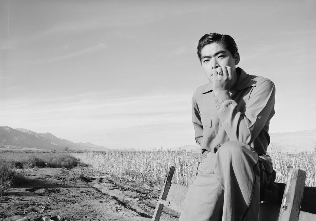 A young man leaning against a wooden fence in Manzanar. His arm is propped up on one of this legs and he is resting his chin on his hand with a pensive look.