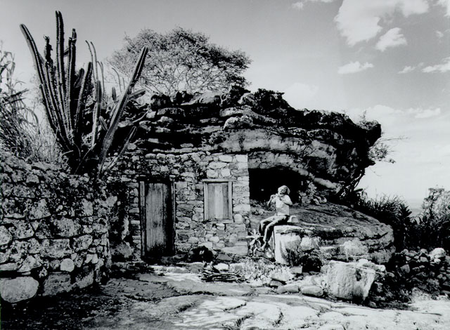 Joaquim Riachão, o "cantador do sertão", sentado sobre uma mureta de pedra, em frente à casa, tocando sua flauta.