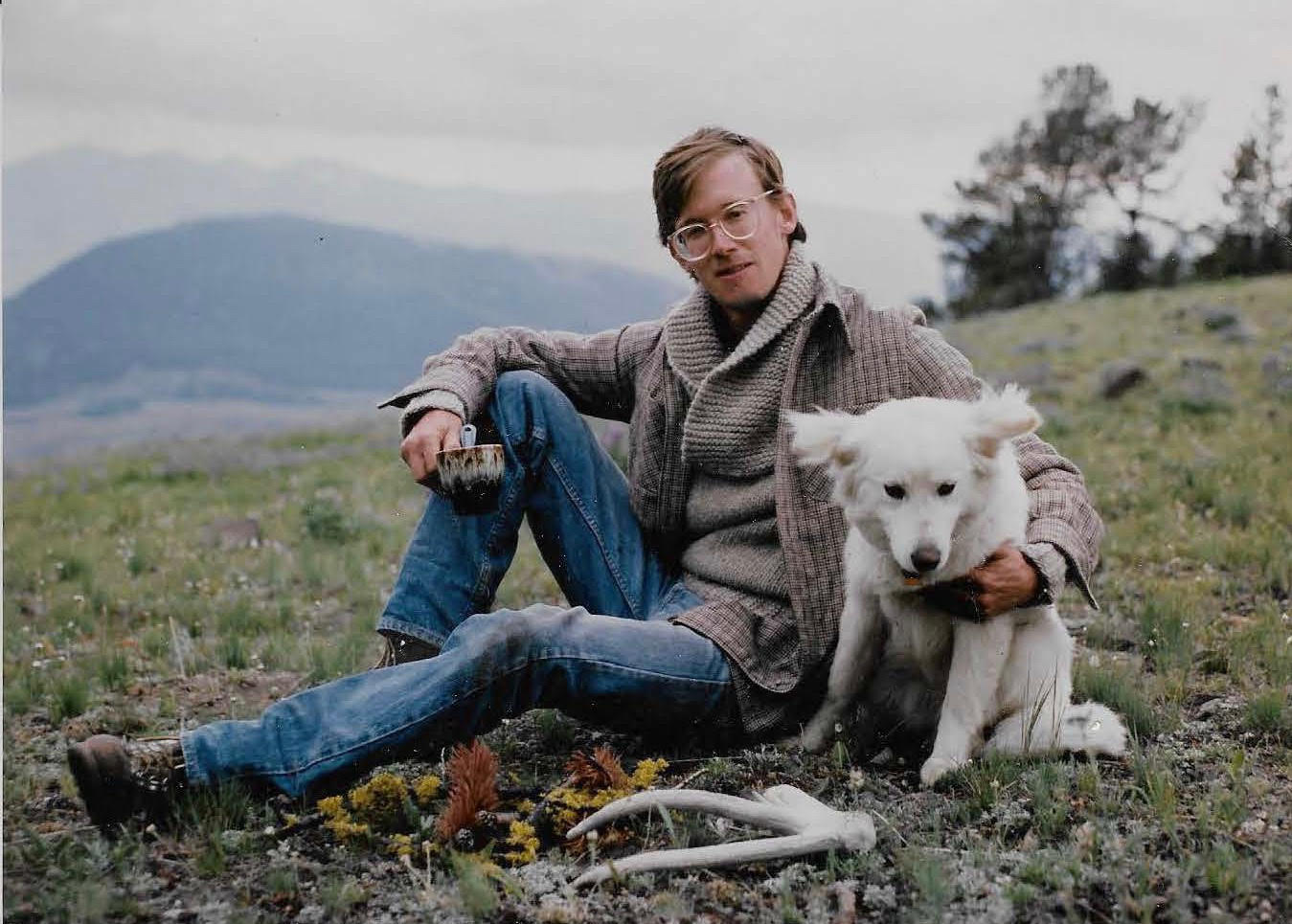 John sitting on a hill with his dog Pie, wearing wide-rimmed glasses, blue jeans, and boots.