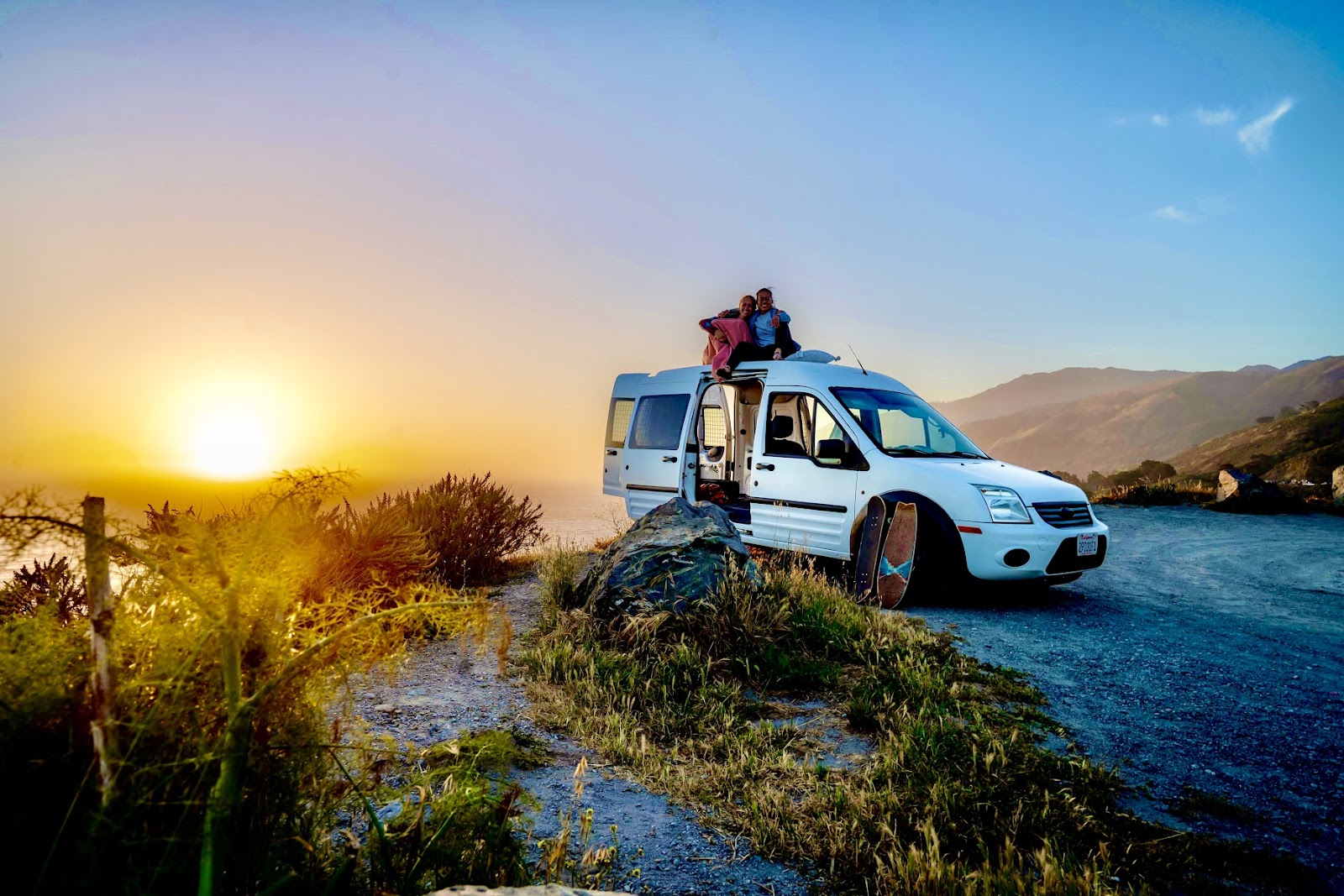 Huemer and Zhong sit atop the van against a scenic landscape and setting sun