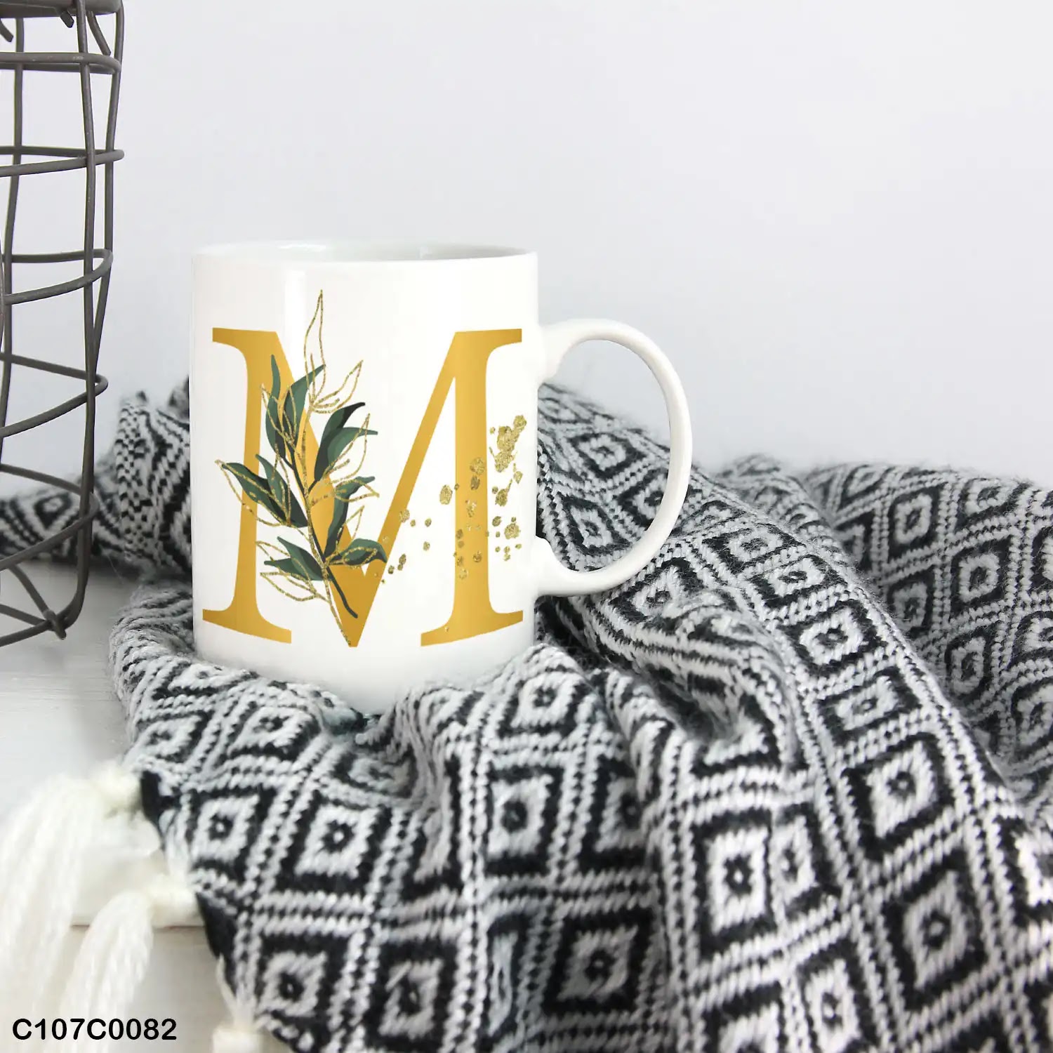 A white mug (cup) printed with gold Letter "M" and small green branch