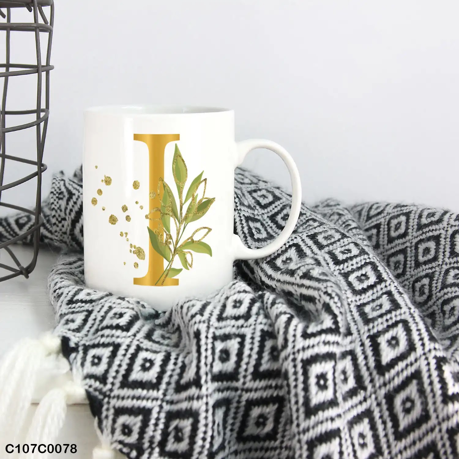 A white mug (cup) printed with gold Letter "I" and small green branch