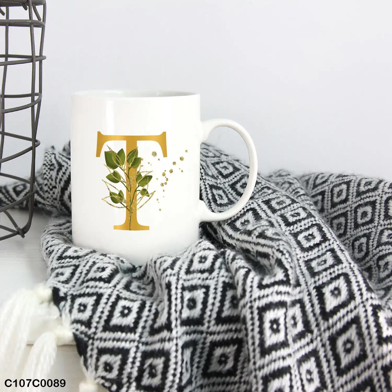 A white mug (cup) printed with gold Letter "T"and small green branch