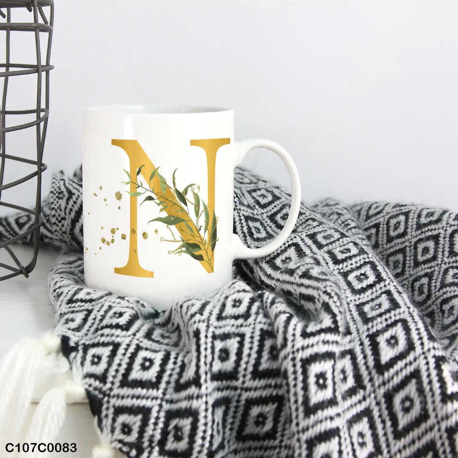 A white mug (cup) printed with gold Letter "N" and small green branch