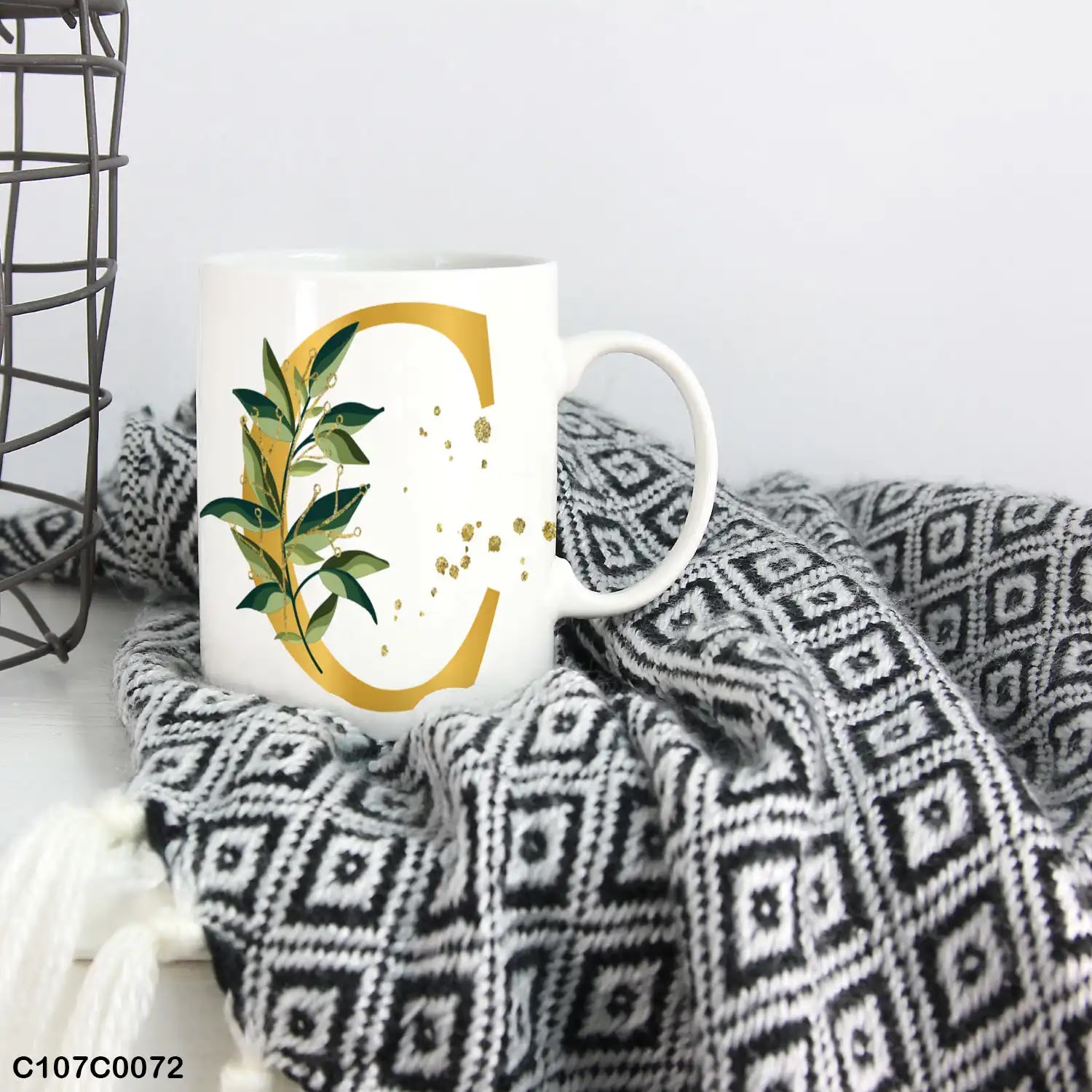A white mug (cup) printed with gold Letter "C" and small green branch