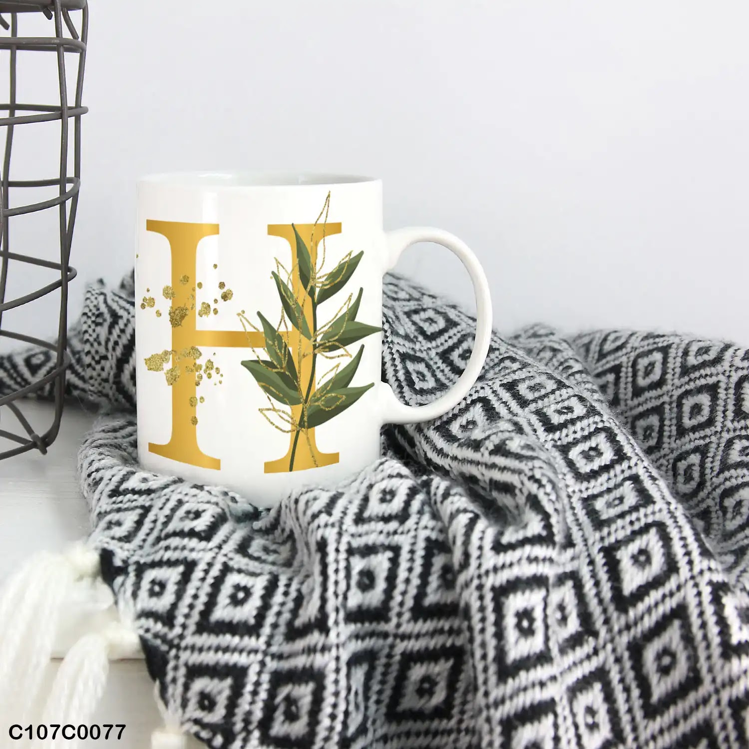 A white mug (cup) printed with gold Letter "H" and small green branch