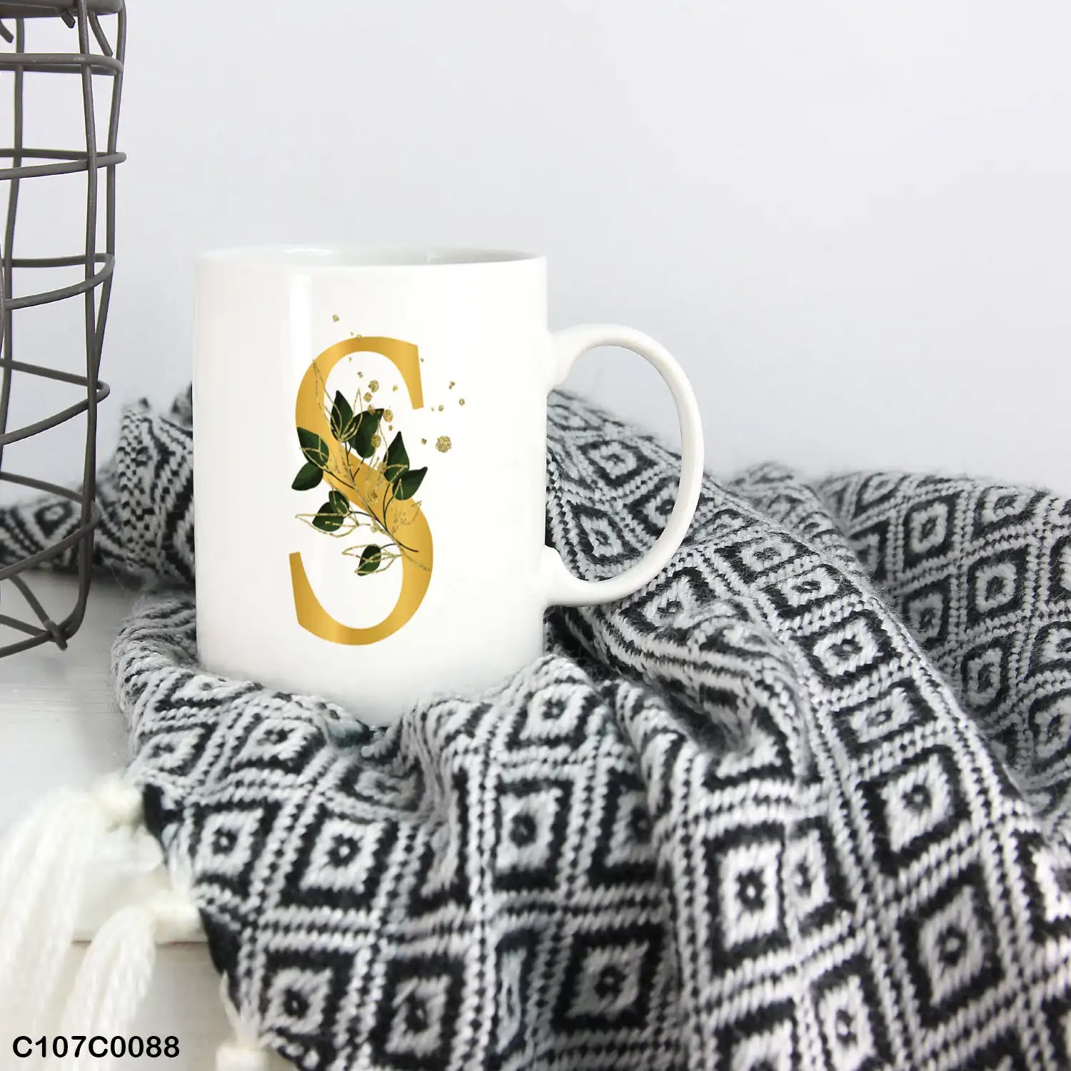 A white mug (cup) printed with gold Letter "S" and small green branch
