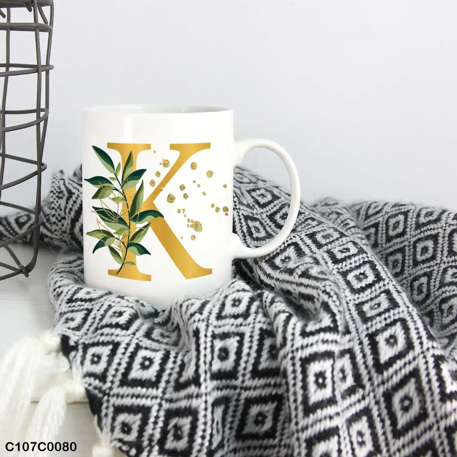 A white mug (cup) printed with gold Letter "K" and small green branch