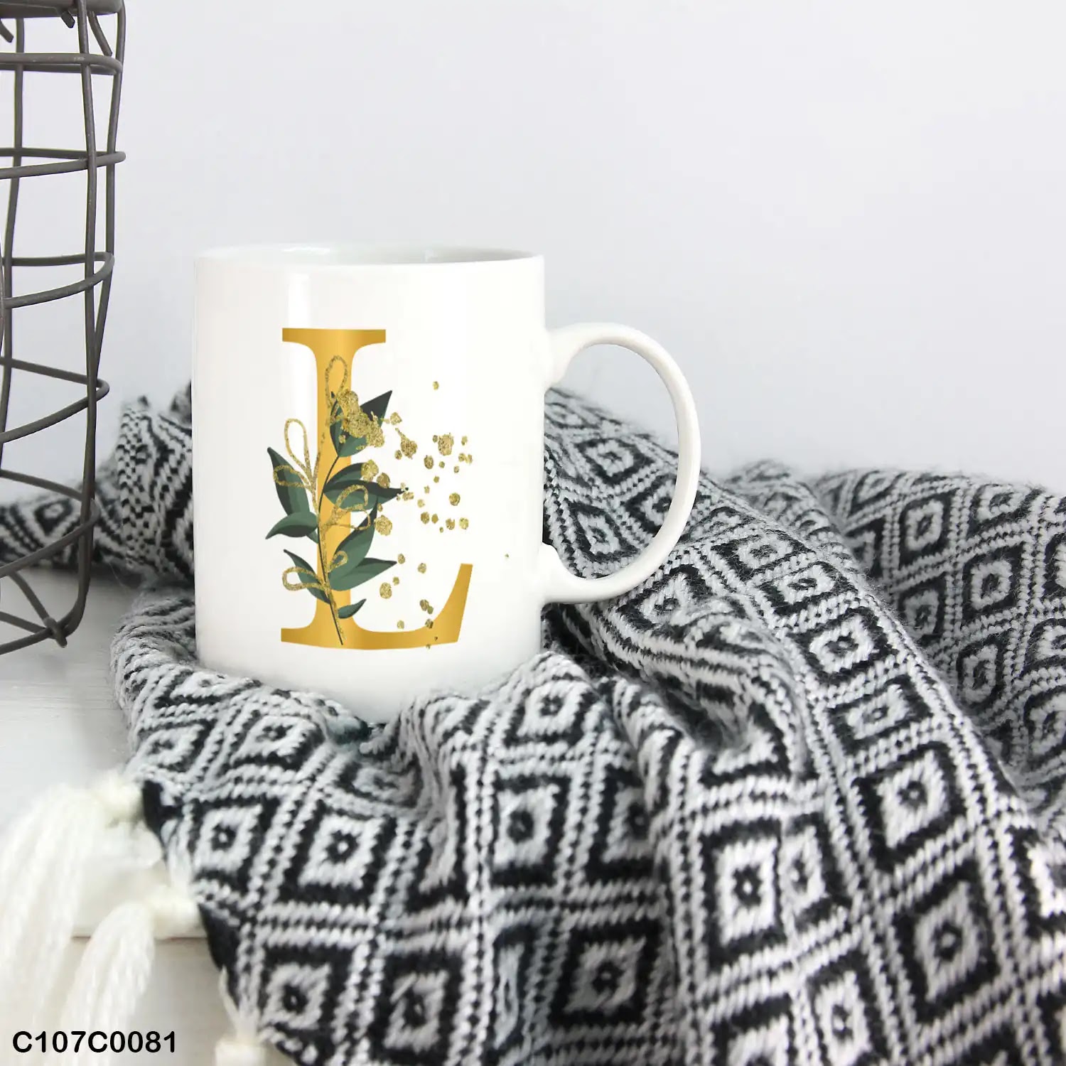 A white mug (cup) printed with gold Letter "L" and small green branch