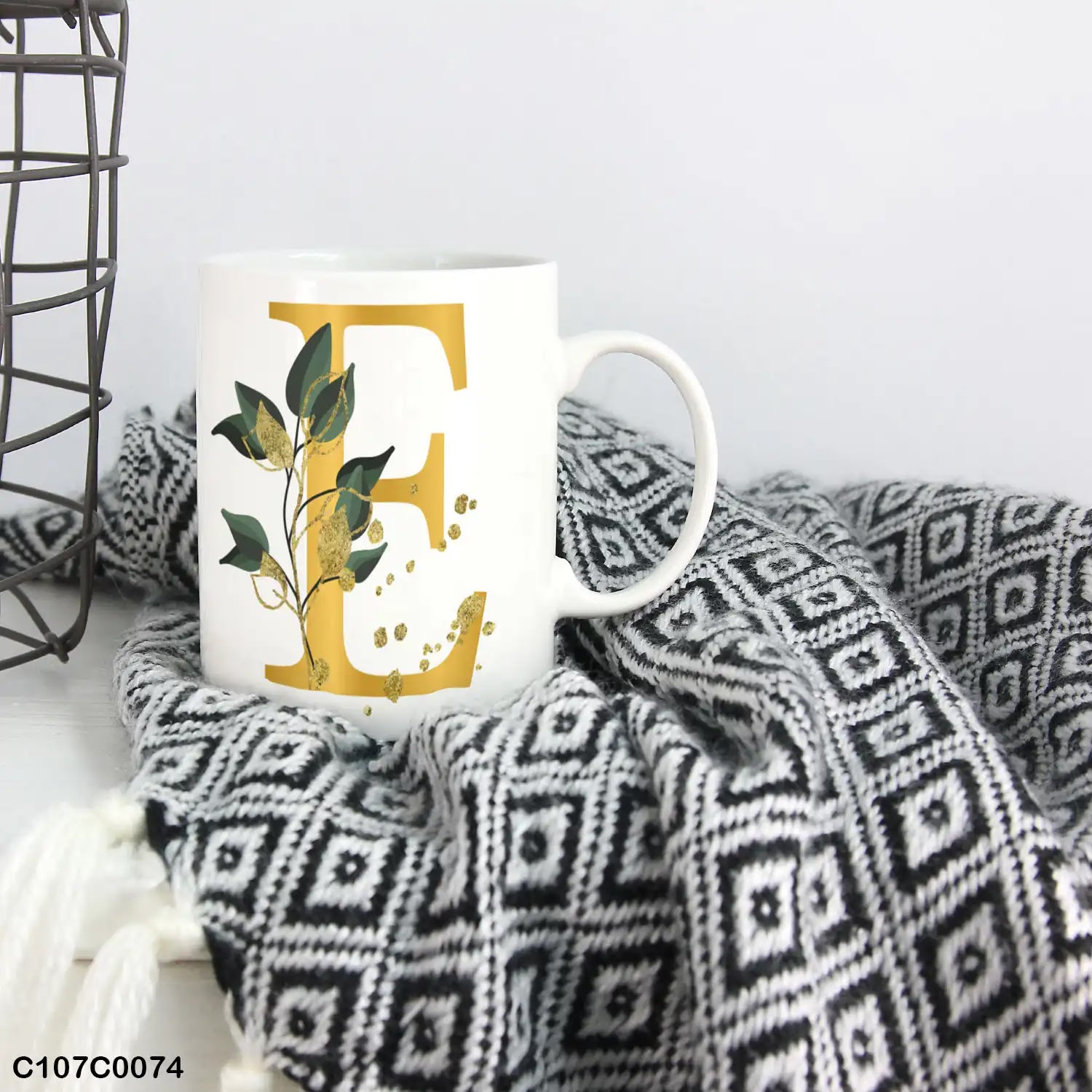 A white mug (cup) printed with gold Letter "E" and small green branch