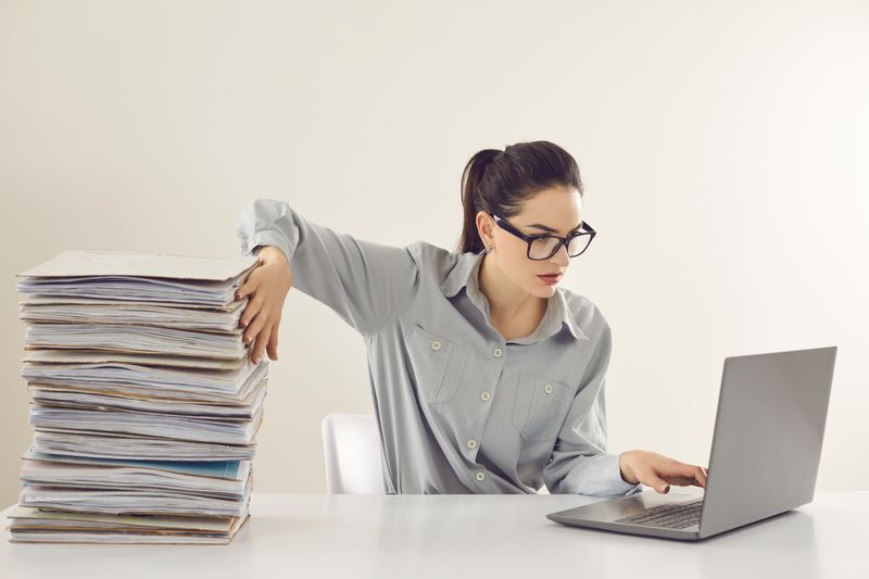 bookkeeping accounting - A woman in a professional outfit with stacked files on her right side and a laptop on her left.