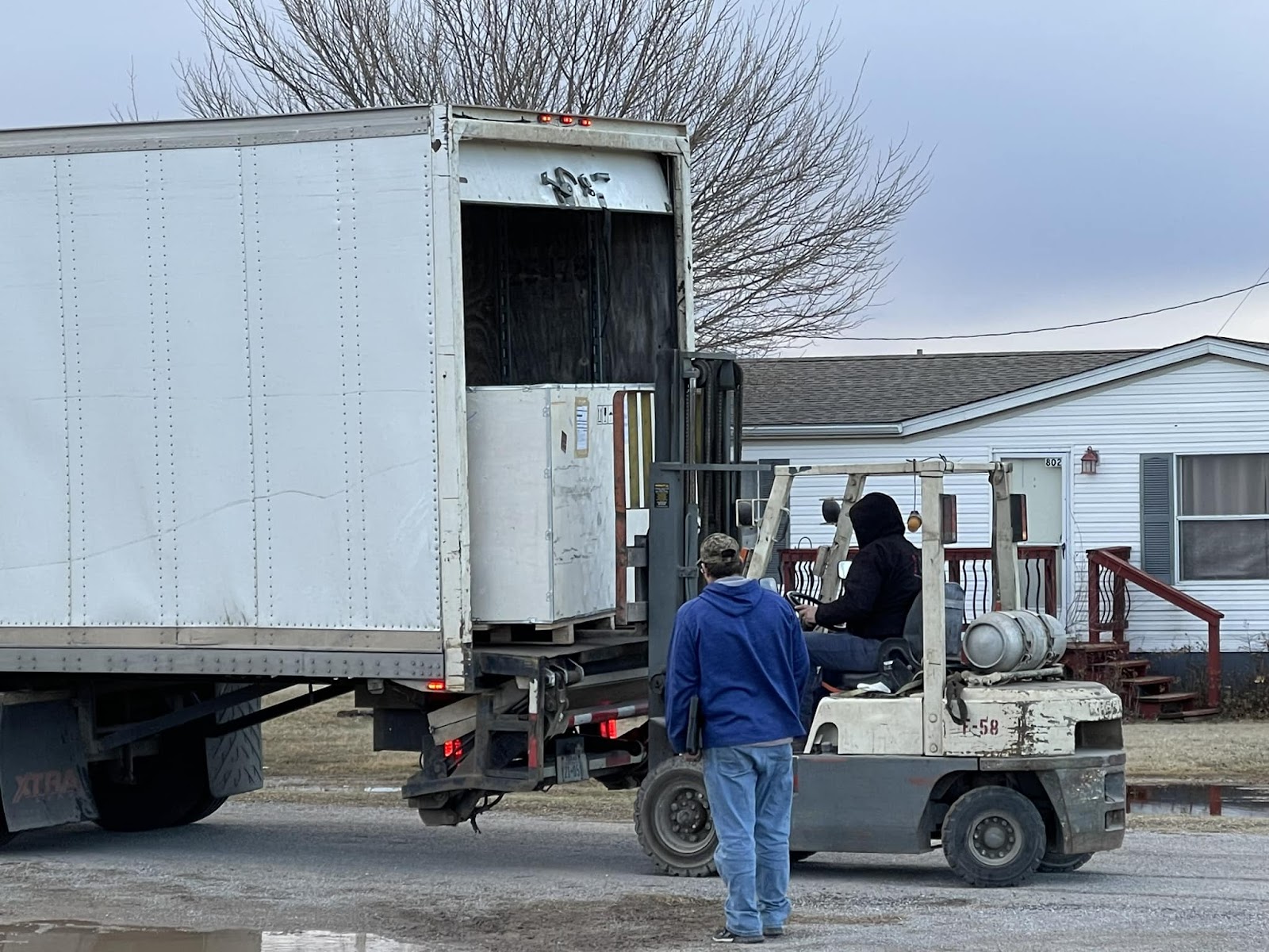 A forklift operator unloads a large industrial crate from the back of a semi-truck trailer