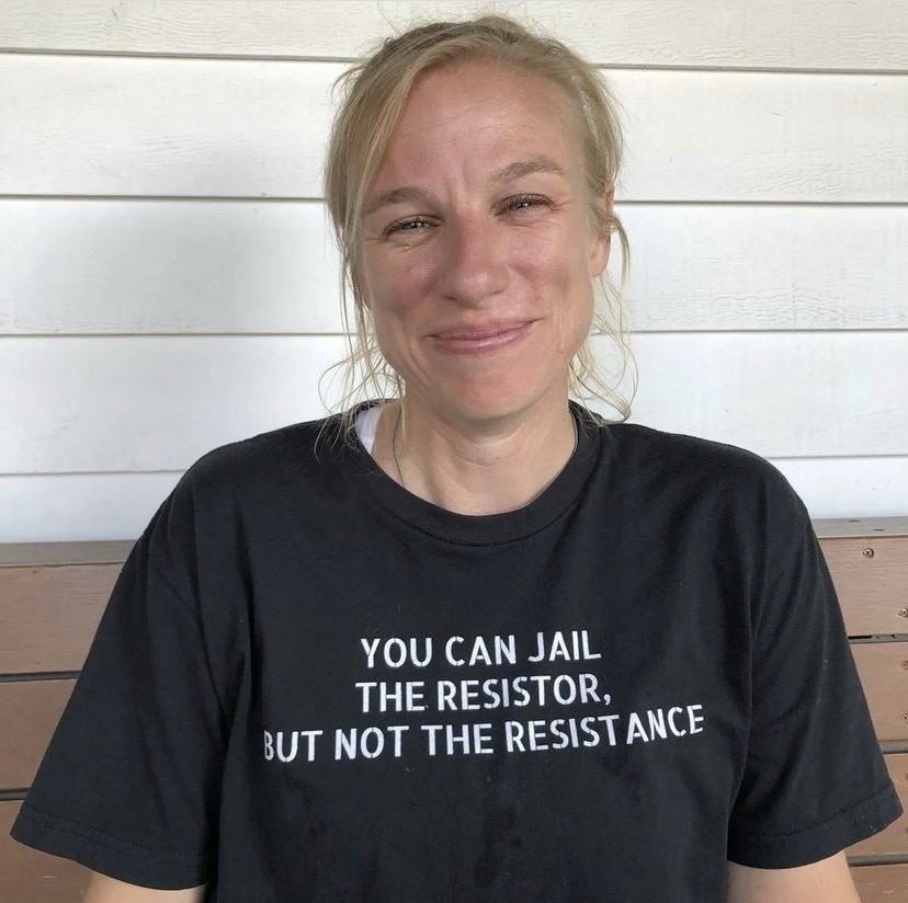 A middle aged blonde woman smiles to camera. She wears a T-shirt that says 'you can jail the resistor but not the resistance'.