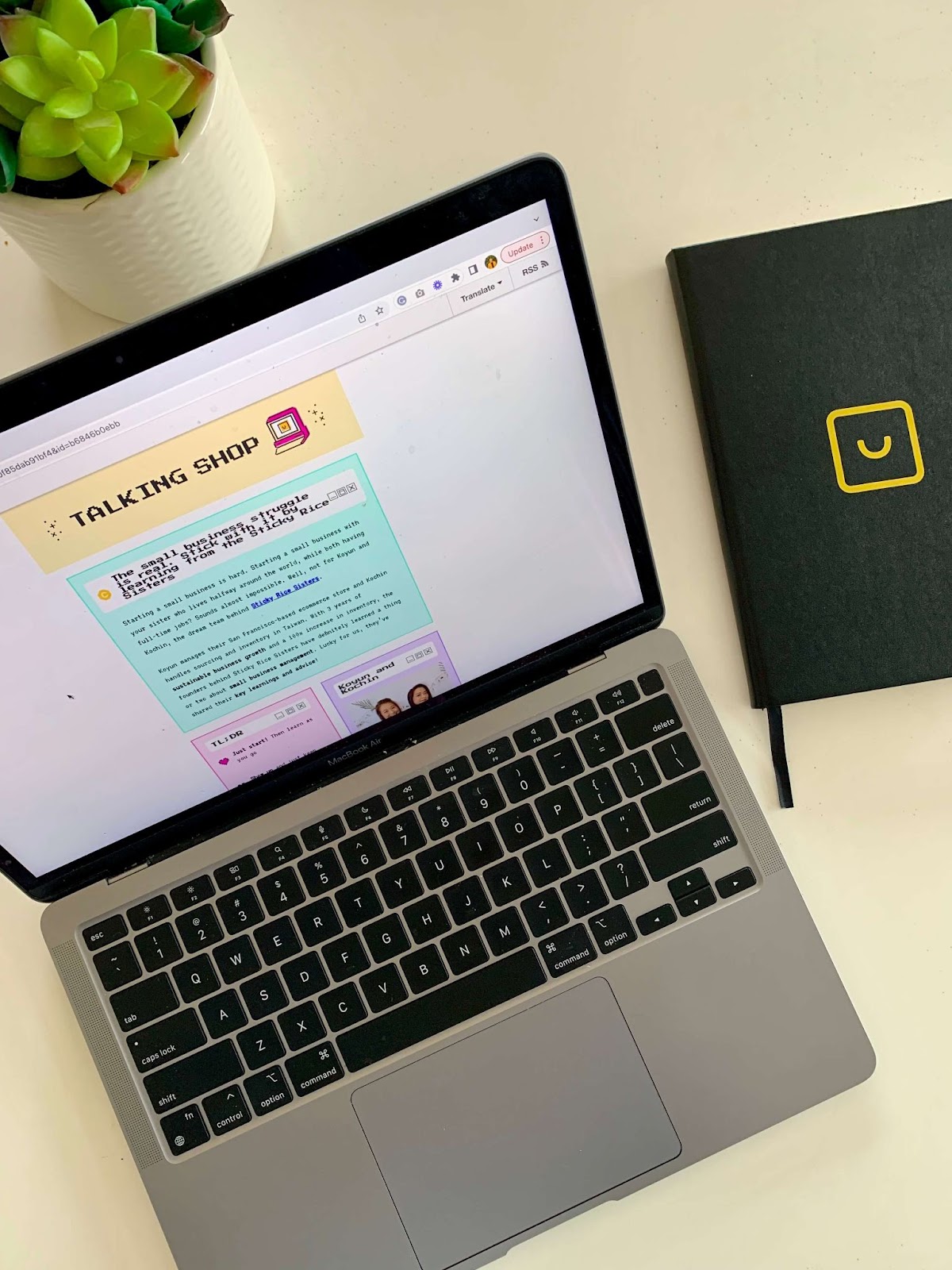 Talking Shop Smile Newsletter–An image of a MacBook on a white table open to the Talking Shop newsletter, which is an 8-bit style newsletter in bright colors. There is a black notebook with the yellow smile logo on it and a succulent plant on the table. 