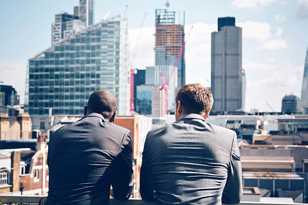 Two businessmen looking at city scape Back view of two businessmen - caucasian and afro american - standing outdoor and looking at city scape.  Investing in Real Estate stock pictures, royalty-free photos & images