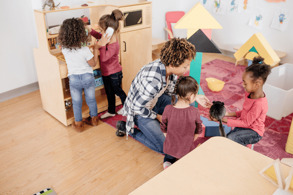 Teacher sitting on the ground with two preschool age girls. One girl is holding a brown kitten while the teacher is leaning towards her. In the background, two other preschool age girls are playing at the sink of a wooden toy kitchen.