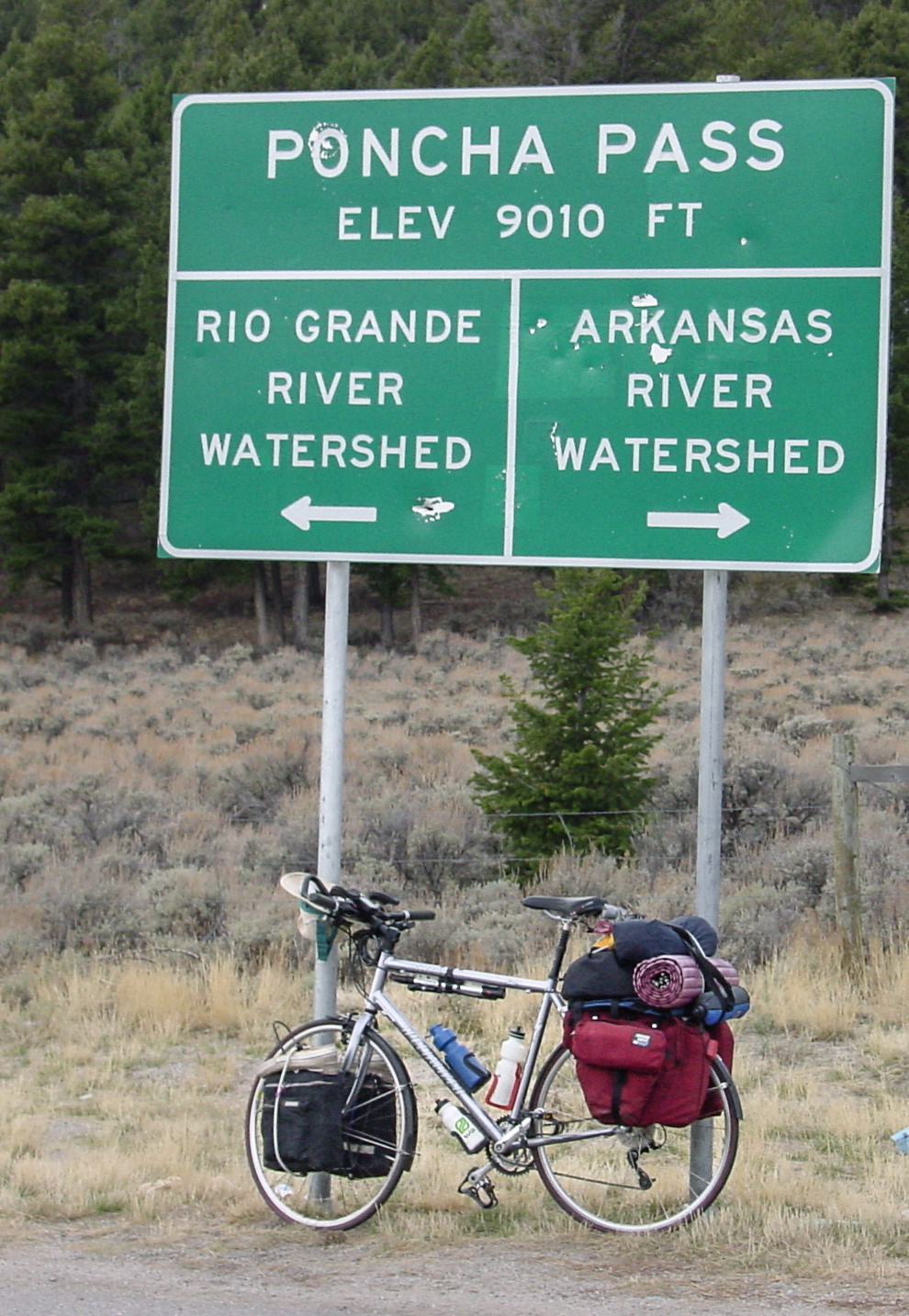 Bicycle leans on a sign that says:PONCHA PASS ELEV 9,010 FT. Arrow pointing left says: RIO GRANDE RIVER WATERSHED; Arrow pointing right says: ARKANSAS RIVER WATERSHED