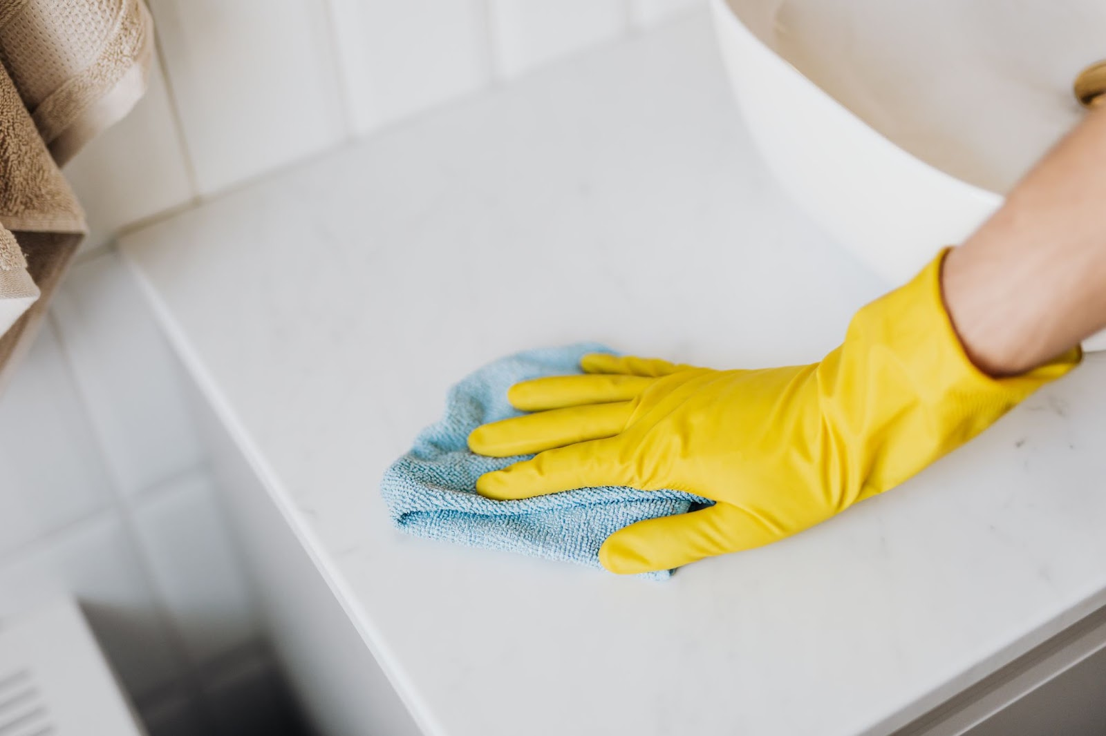 Hand with yellow rubber glove wiping a white countertop with a blue microfibre cloth.