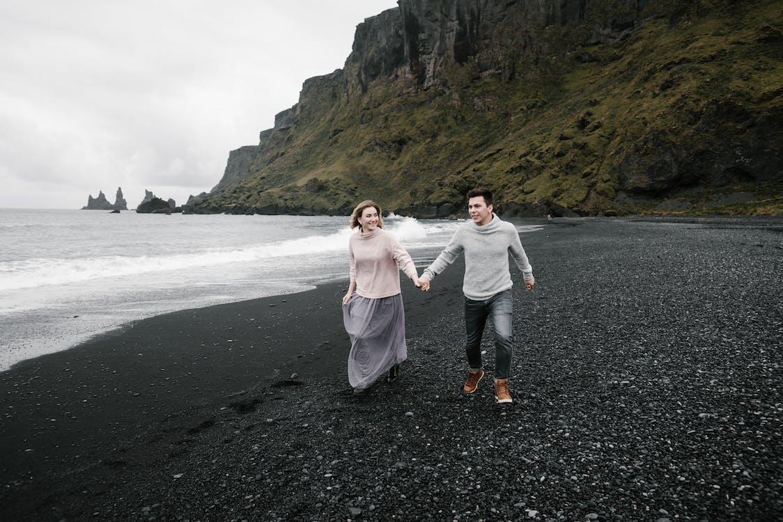 Free Happy loving couple holding hands and exploring magnificent black gravel beach while enjoying weekend at cliffy seacoast Stock Photo