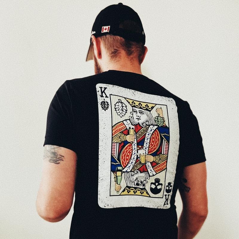 A man with his back to the camera, wearing a black T-shirt with a graphic on the back.  The graphic looks like a King playing card holding a beer and hops. 