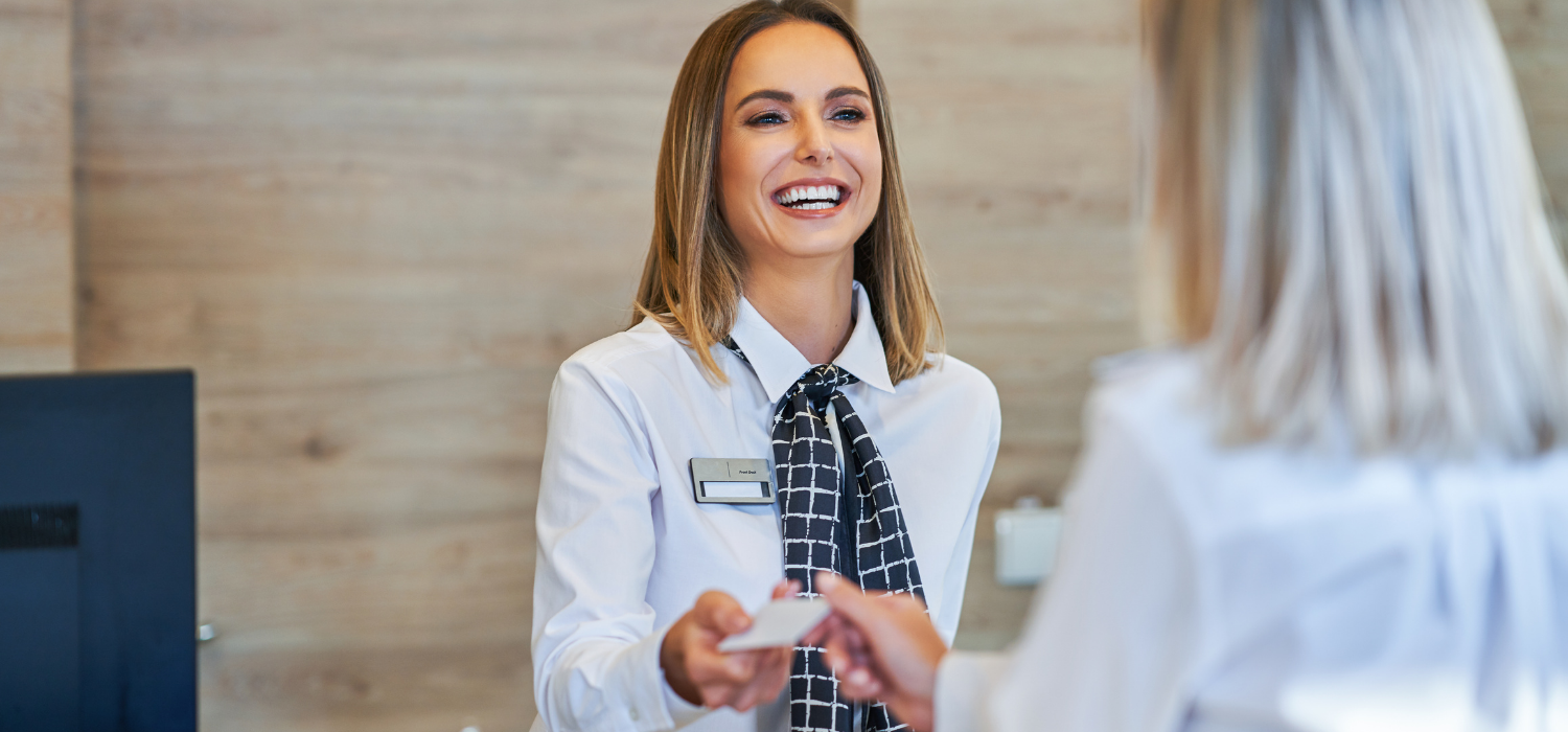 a hotel receptionist in a hotel uniform with a name tag attached happily explaining how hotel booking websites work to a blonde female guest. 