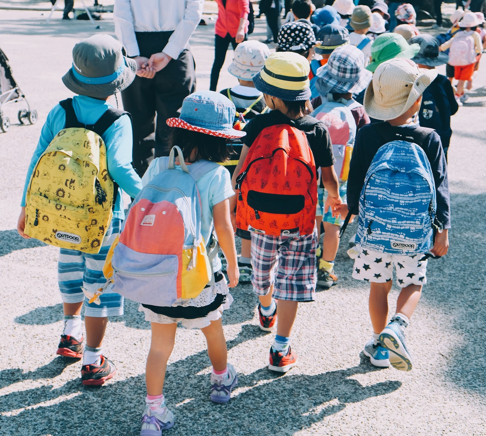 A single file line of elementary students with backpacks travels alongside several teachers.